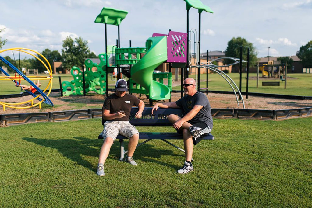 Two men sitting on a park bench with kids playing in the background.